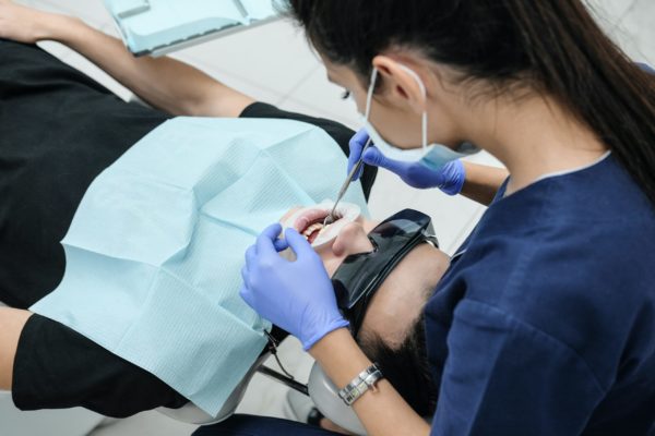 A photo of a dentist with long dark hair taking care of a patient wearing the blue napkin around their neck.