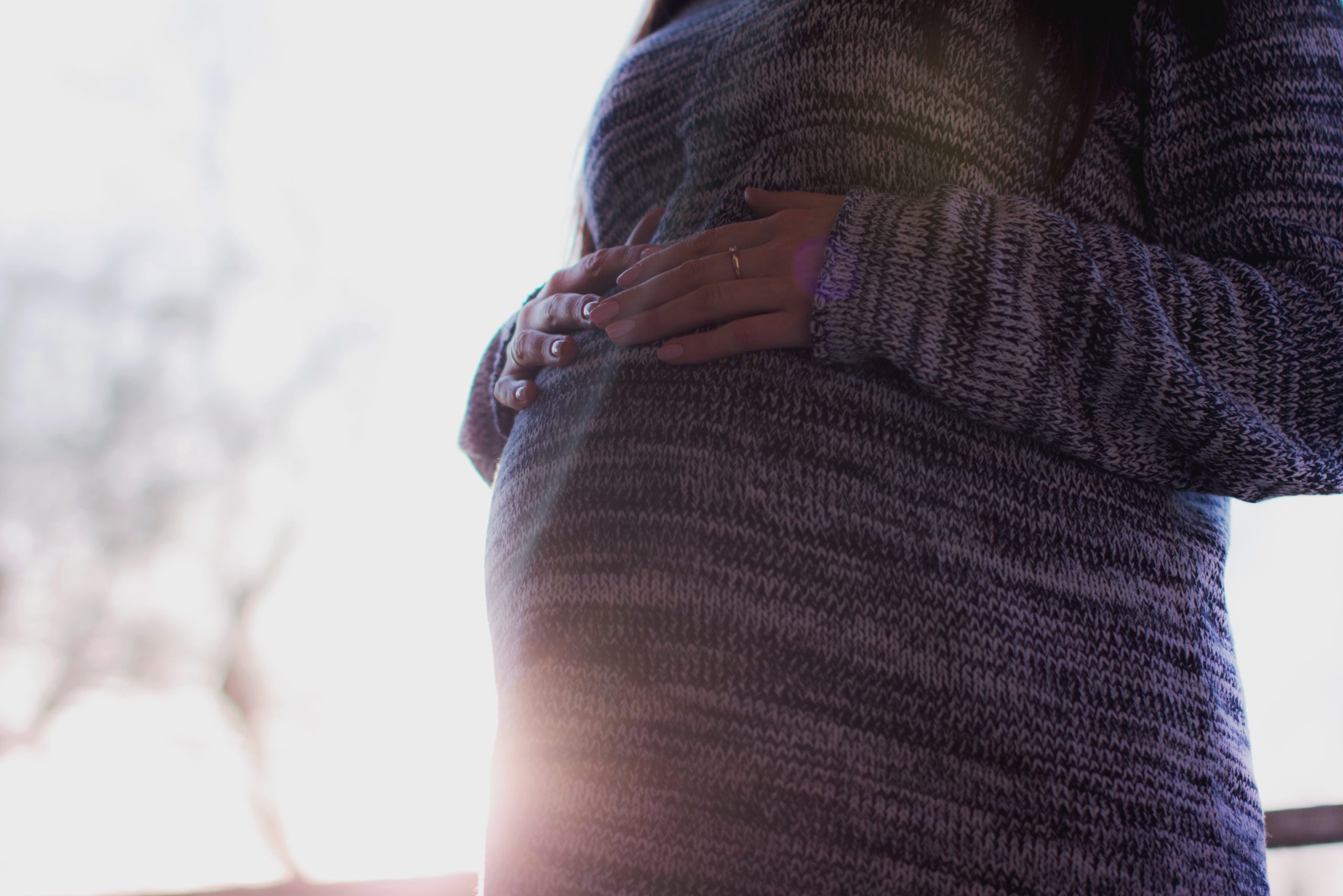 A pregnant woman placing both of her hands on her belly as she's backlit by the sunset.