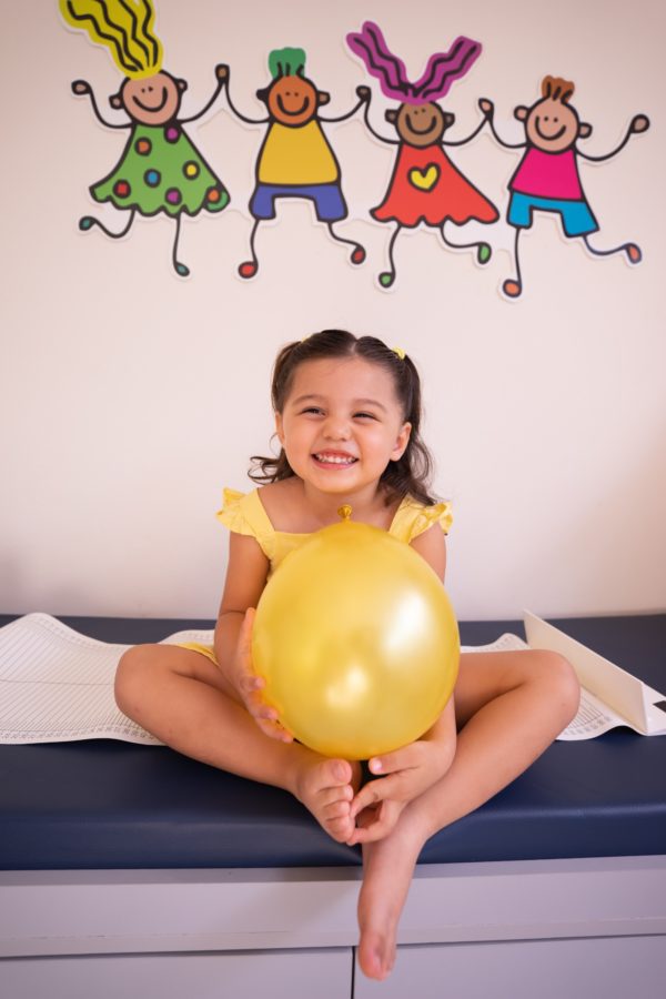 A small girl wearing bright yellow at the pediatricians holding a bright yellow balloon.